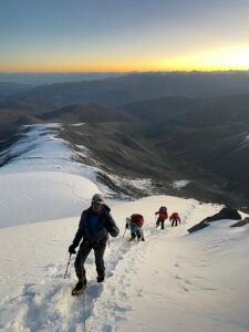 Four climbers roped together ascending a snowy mountain slope during sunrise or sunset.
